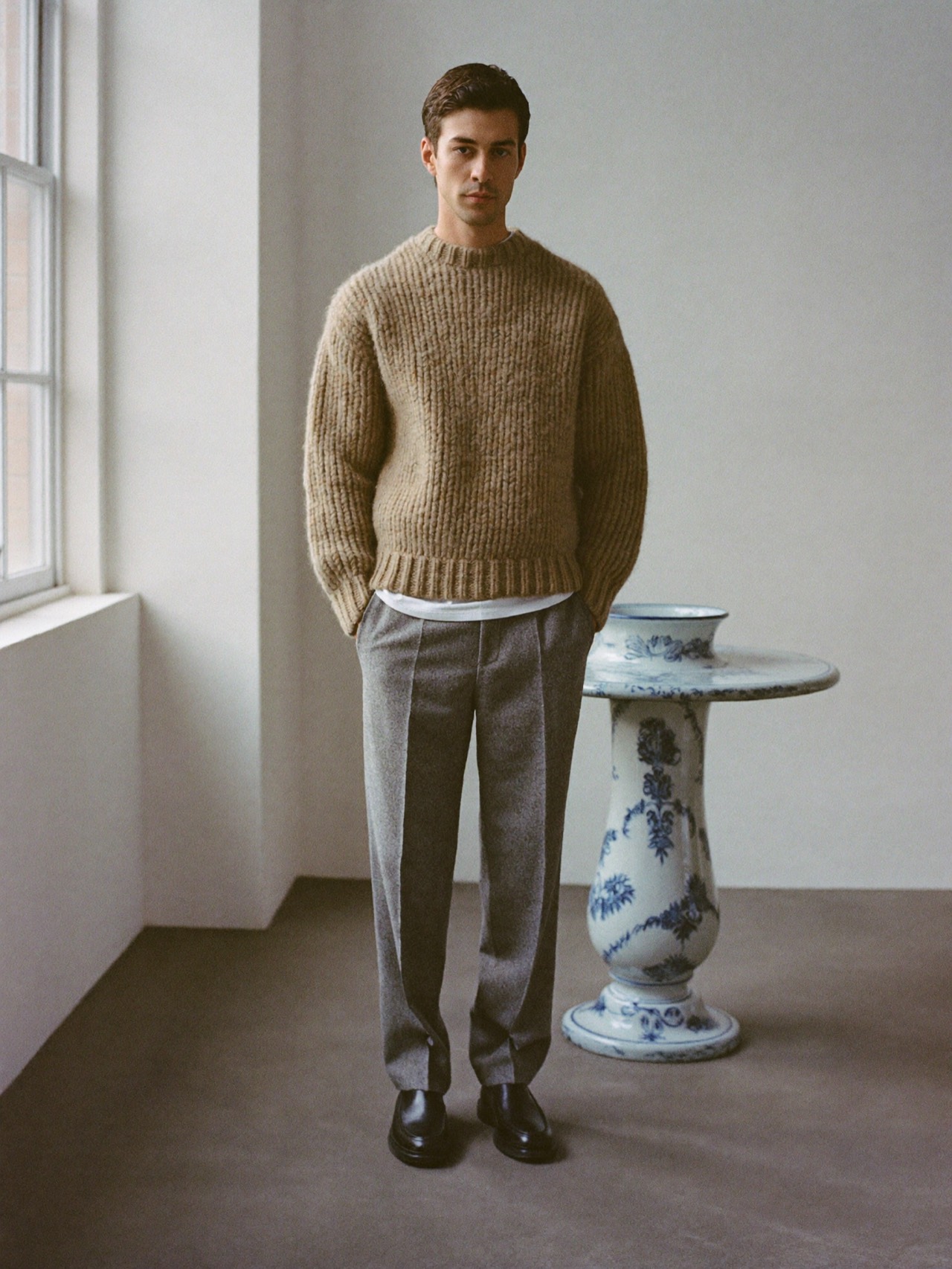 Man in an oatmeal knit sweater and grey wool trousers against a porcelain studio wall